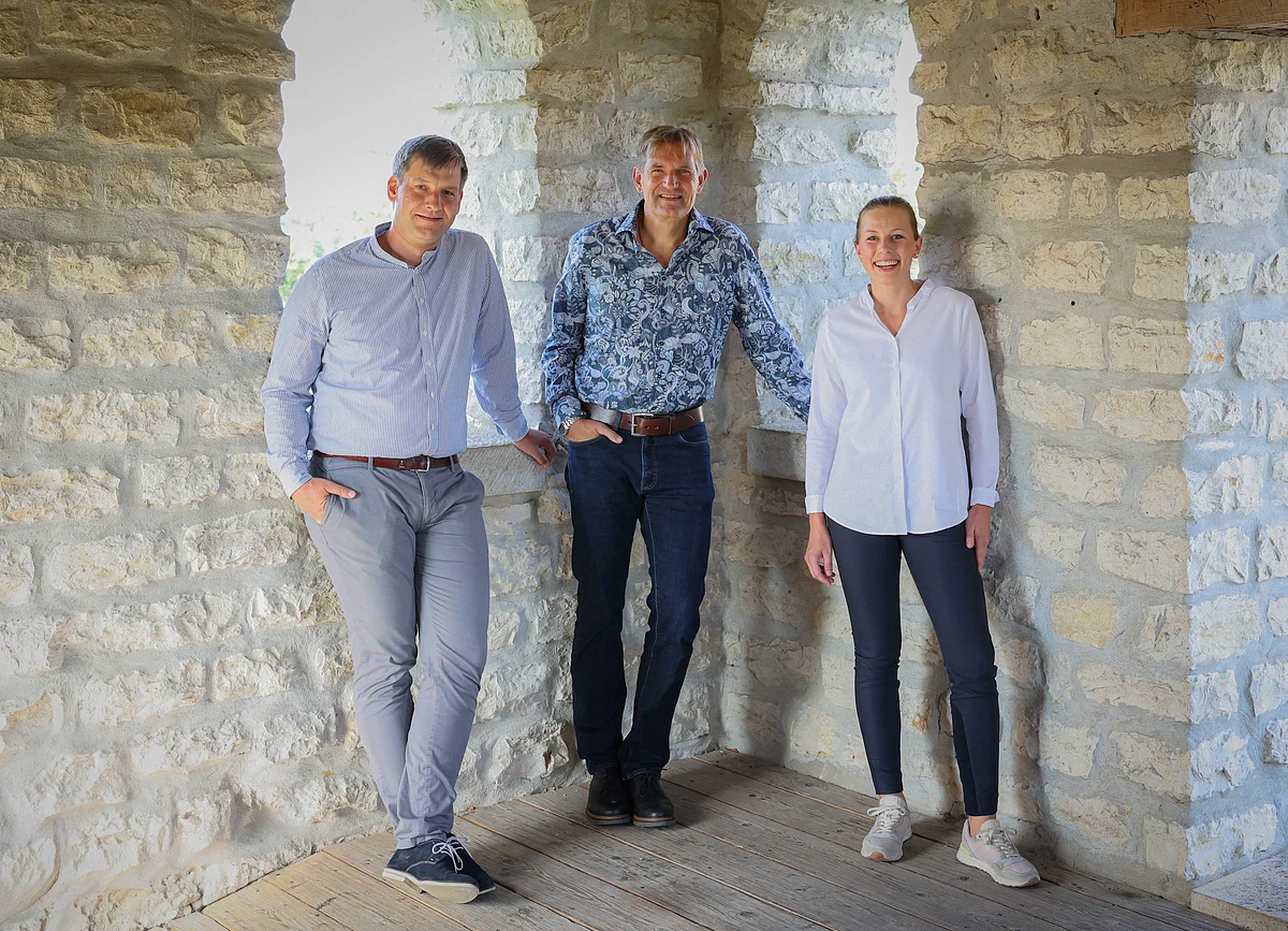 Drei Personen stehen in einem steinernen Raum mit Holzboden und blicken in die Kamera.